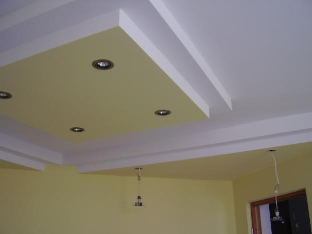 Pomarańczowo-biały sufit podwieszany z zamontowanymi czterema okrągłymi lampami halogenowymi, widoczne kable zwisające z sufitu w innym miejscu pokoju, ściany pomalowane na pomarańczowo.